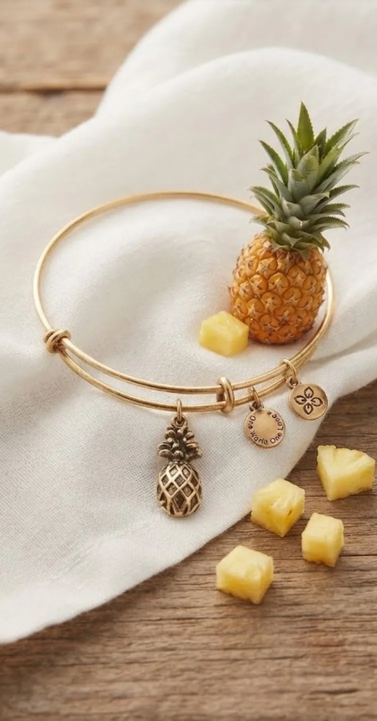 A gold-tone pineapple charm bangle on white linen with fresh pineapple