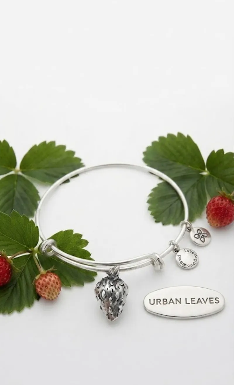 A silver-tone strawberry bangle among wild strawberries and their leaves, beside the Urban Leaves plaque
